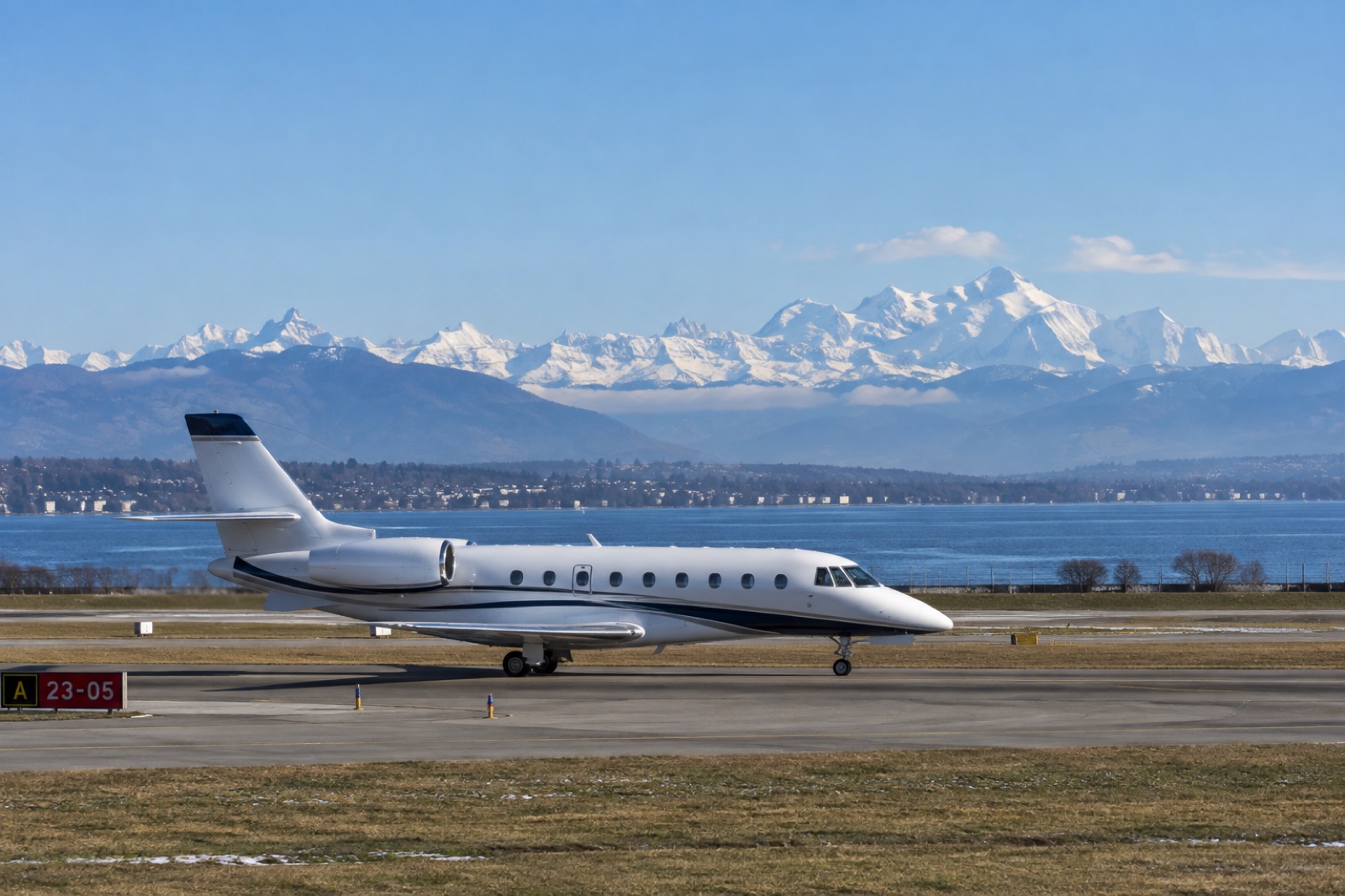 A long-range business jet taxiing at Geneva on a clear winter afternoon, the lake and the snow-covered Alps on the horizon
