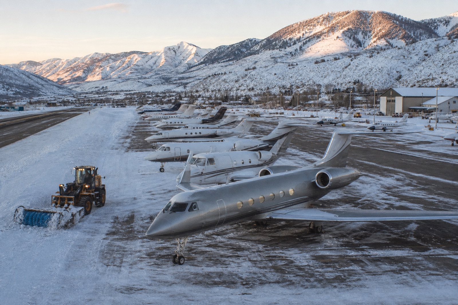A crowded, snow-covered general aviation ramp at a mountain airport, a tug clearing snow between parked business jets