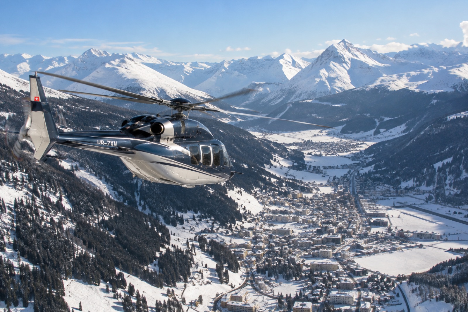 A twin-engine executive helicopter flying up the snow-covered valley toward Davos on a sunny January afternoon